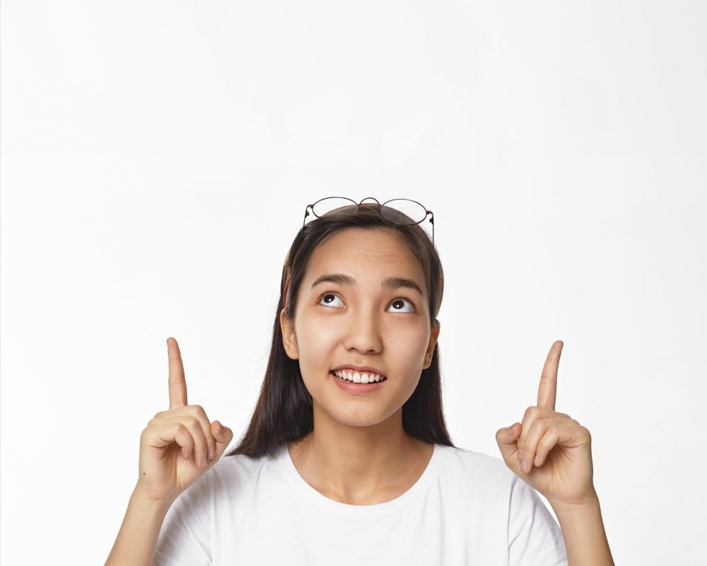 Look at this. Picture of happy emotional young Chinese woman in casual clothes looking up and pointing both index fingers. Indicating blank white studio wall with copyspace for your advertisement