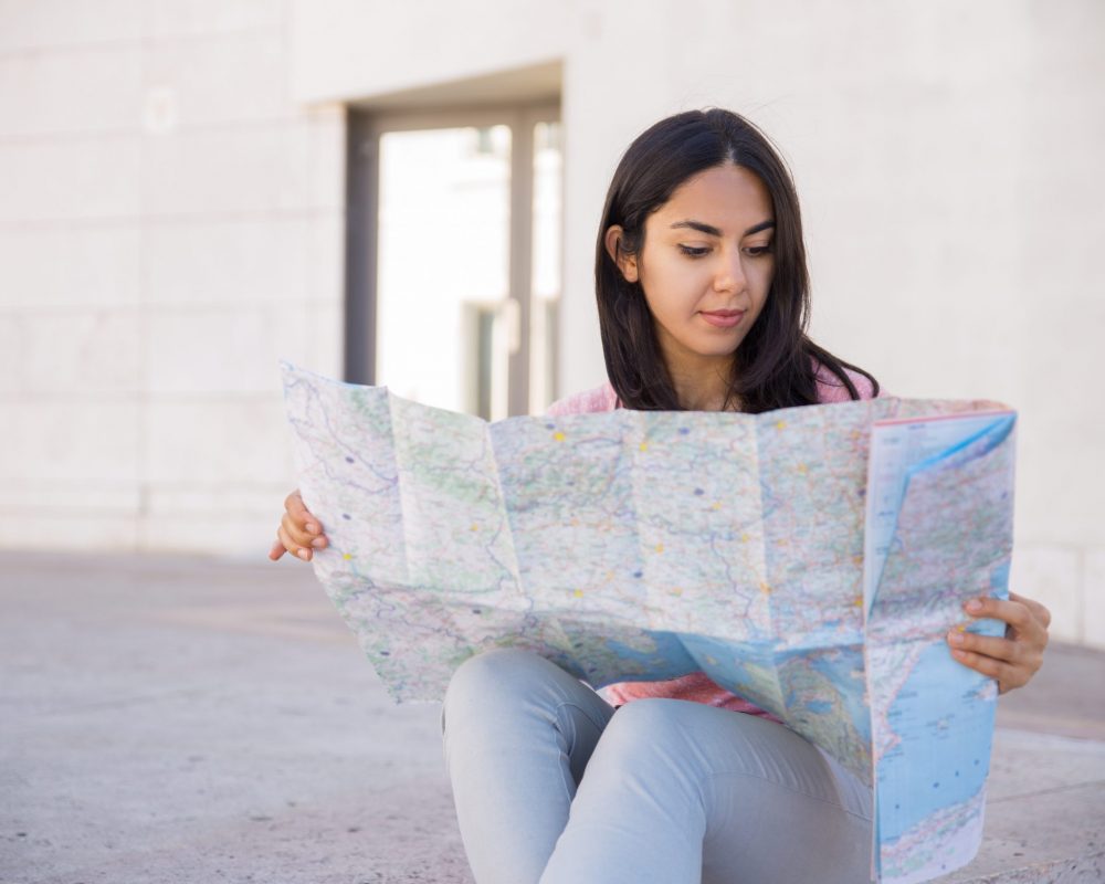 Focused young woman studying paper map outdoors. Pretty lady sitting with building wall in background. Vacation and tourism concept. Front view.